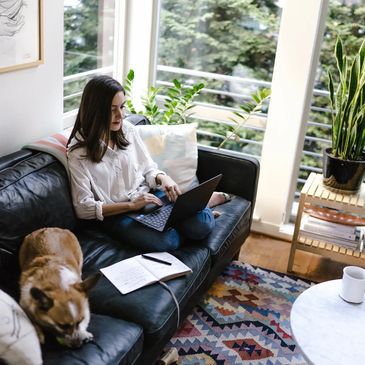 Relaxed woman works on  a media project on her laptop while sitting on a couch beside a small dog.