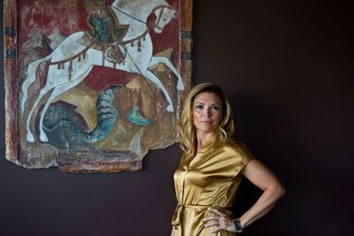 Woman in a gold dress poses beside a medieval-style painting of a knight on a white horse.