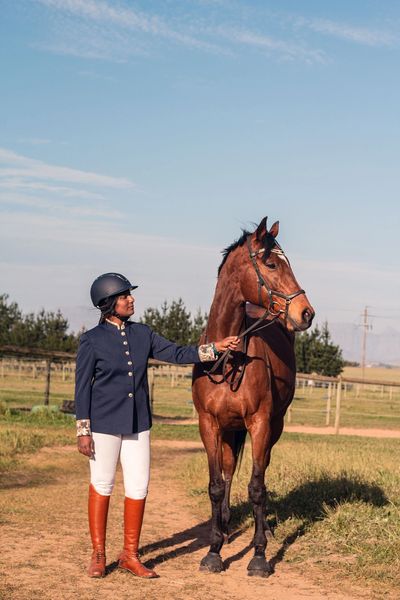 A rider in equestrian attire stands beside a brown horse on a sunny day.