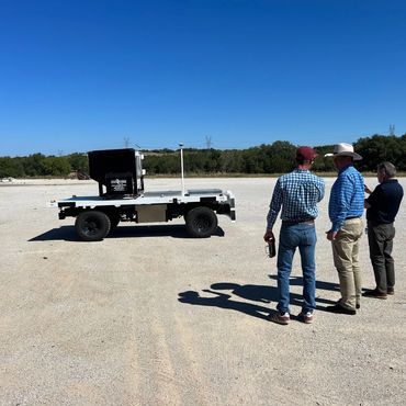 Three men observe a robotic vehicle on a gravel surface under a clear blue sky.