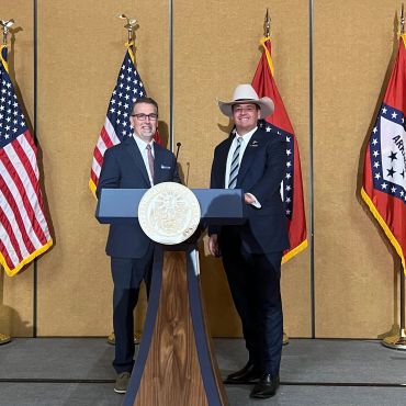 Two men in suits stand behind a podium with American and Arkansas flags.