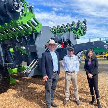 Three people posing in front of large agricultural machinery under a partly cloudy sky.