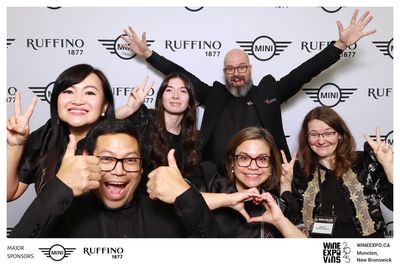 Group of six cheerful people posing with thumbs up, peace signs, and a heart gesture at Wine Expo 2025.