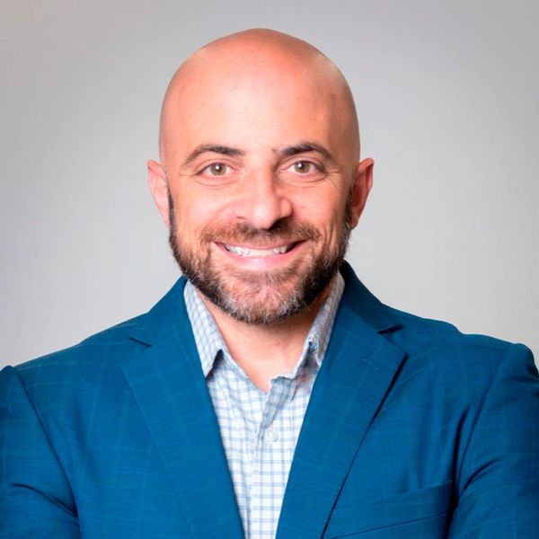 Smiling bald man in a blue blazer and checkered shirt against a light background.