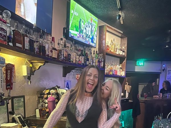Two women happily posing behind a bar counter.