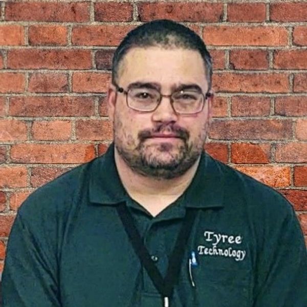 Man with glasses in a Tyree Technology shirt against a brick wall.