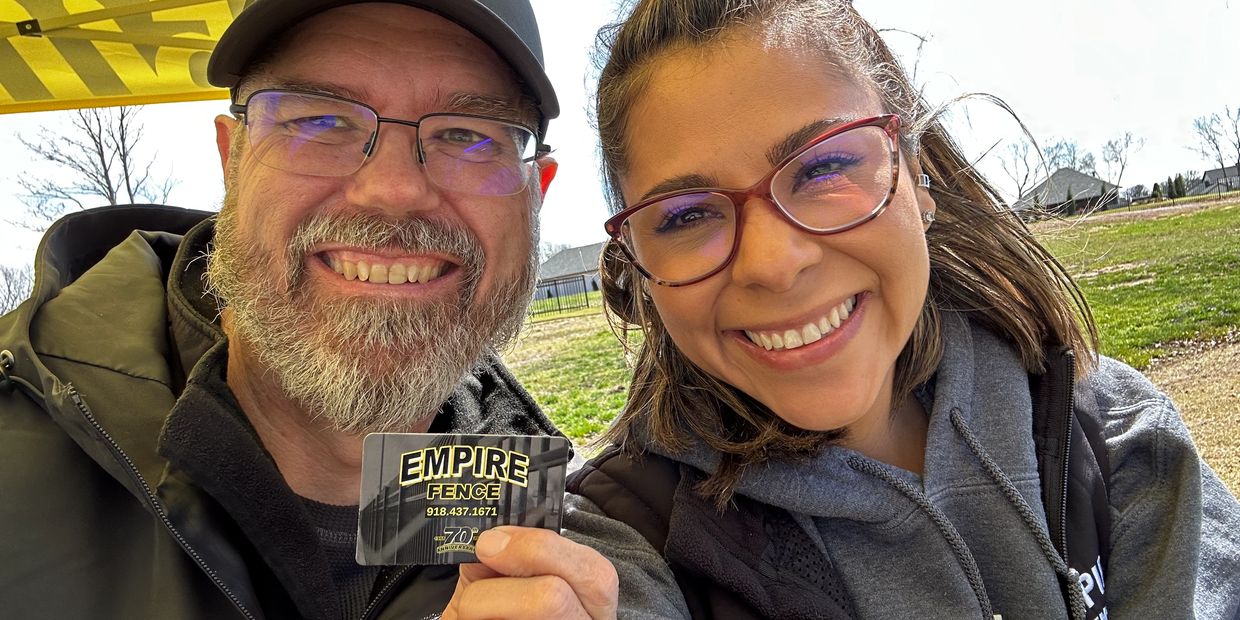Two smiling people holding an Empire Fence card outdoors on a sunny day.