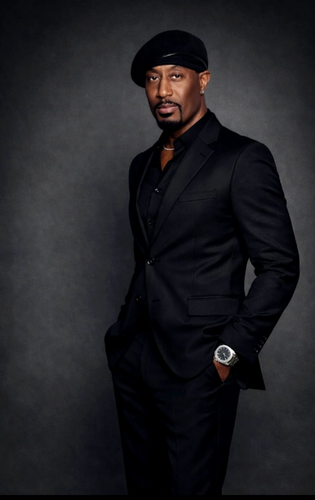 Man in a black suit and beret poses confidently.