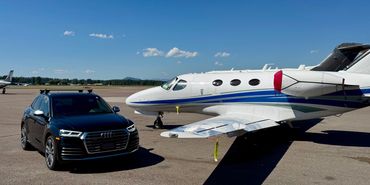 Black Audi SUV parked beside a small private jet on a sunny day.