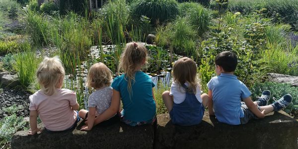 Die Kinder sitzen nebeneinander auf Steinblöcken und schauen auf einen Teich.