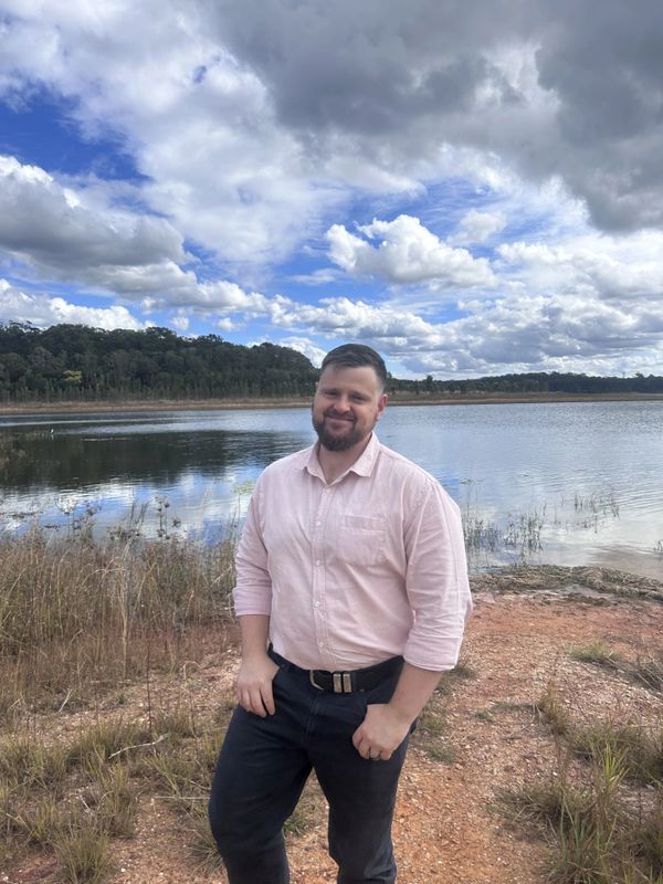 Man in a pink shirt standing by a lakeside under a cloudy sky.