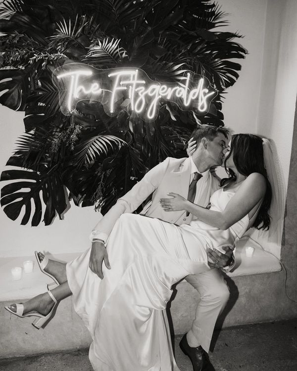 Black and white image of a husband holding his wife in front of a neon sign and a tropical backdrop