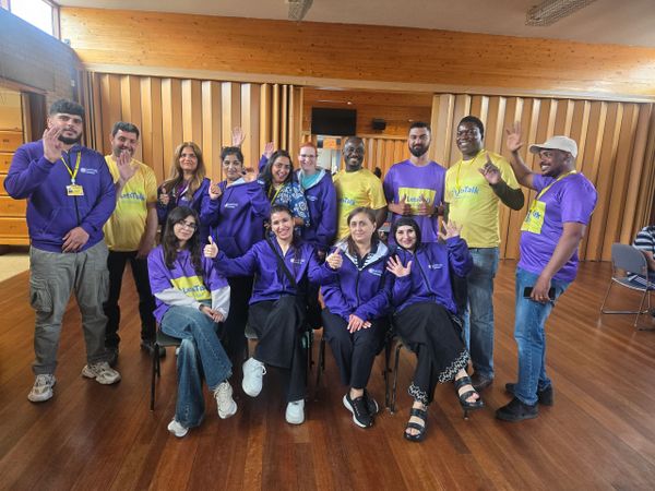 A diverse group of people smiling and waving indoors, wearing yellow and purple shirts.