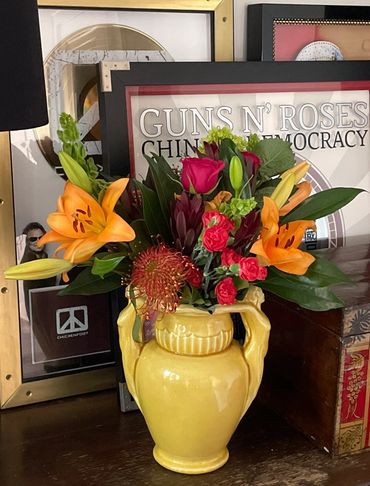 Colorful flowers in a yellow vase with framed music memorabilia in the background.