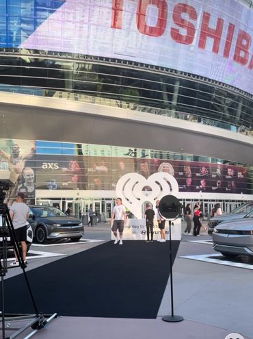 People gather outside a modern venue with cars and a large heart-shaped logo display.