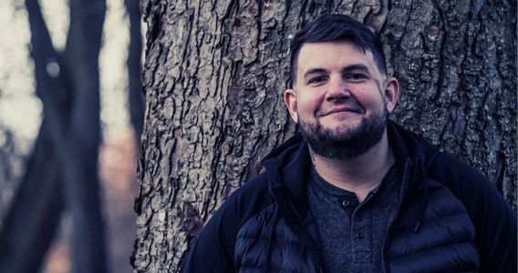 Man with a beard smiling against a large tree trunk outdoors.
