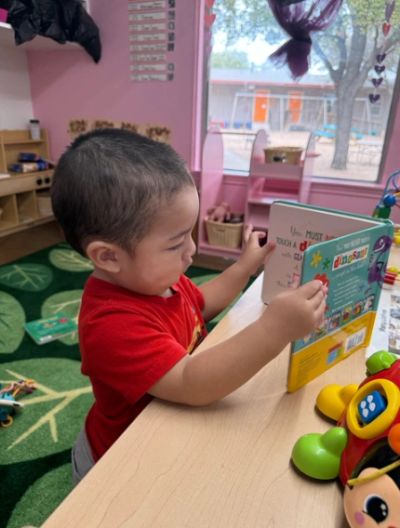Little boy wearing a red shirt, leaning on a bookshelf looking at a book.