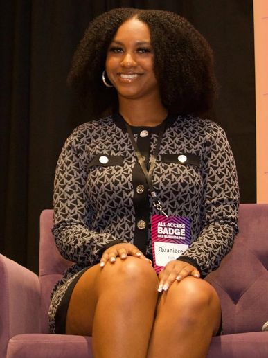 Woman with curly hair smiling, wearing a patterned dress and an event badge.
