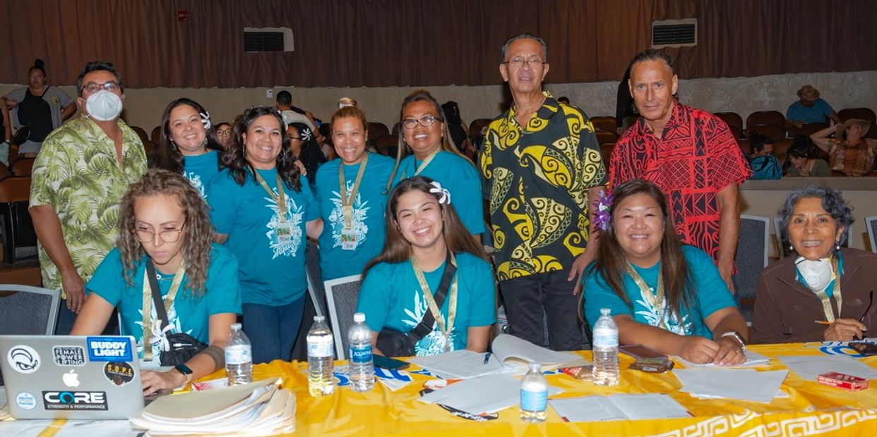Group of people in teal shirts and colorful attire smiling at an event table.