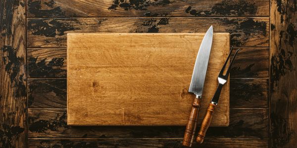 Wooden cutting board with knife and fork on rustic wooden table.