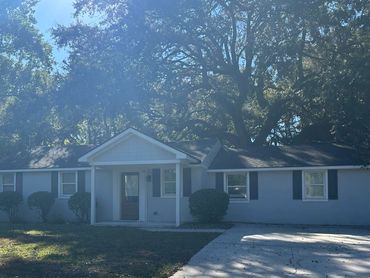 Single-story house with a front porch and driveway under large trees.