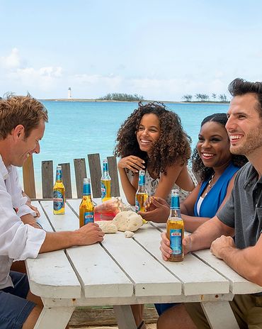 Four friends enjoying beers and laughs by the seaside at a picnic table.