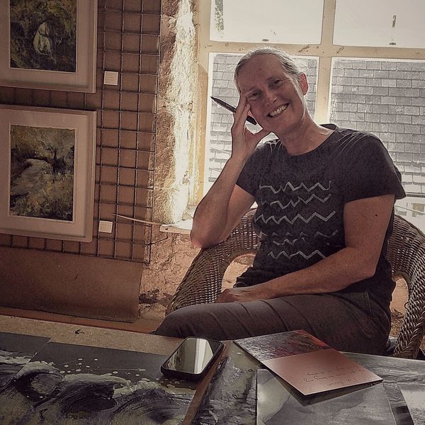 Smiling woman sitting in a wicker chair near a window, surrounded by artwork and art supplies.