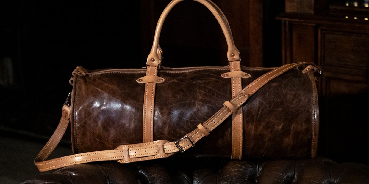 Elegant brown leather duffel bag with tan straps on a leather chair.