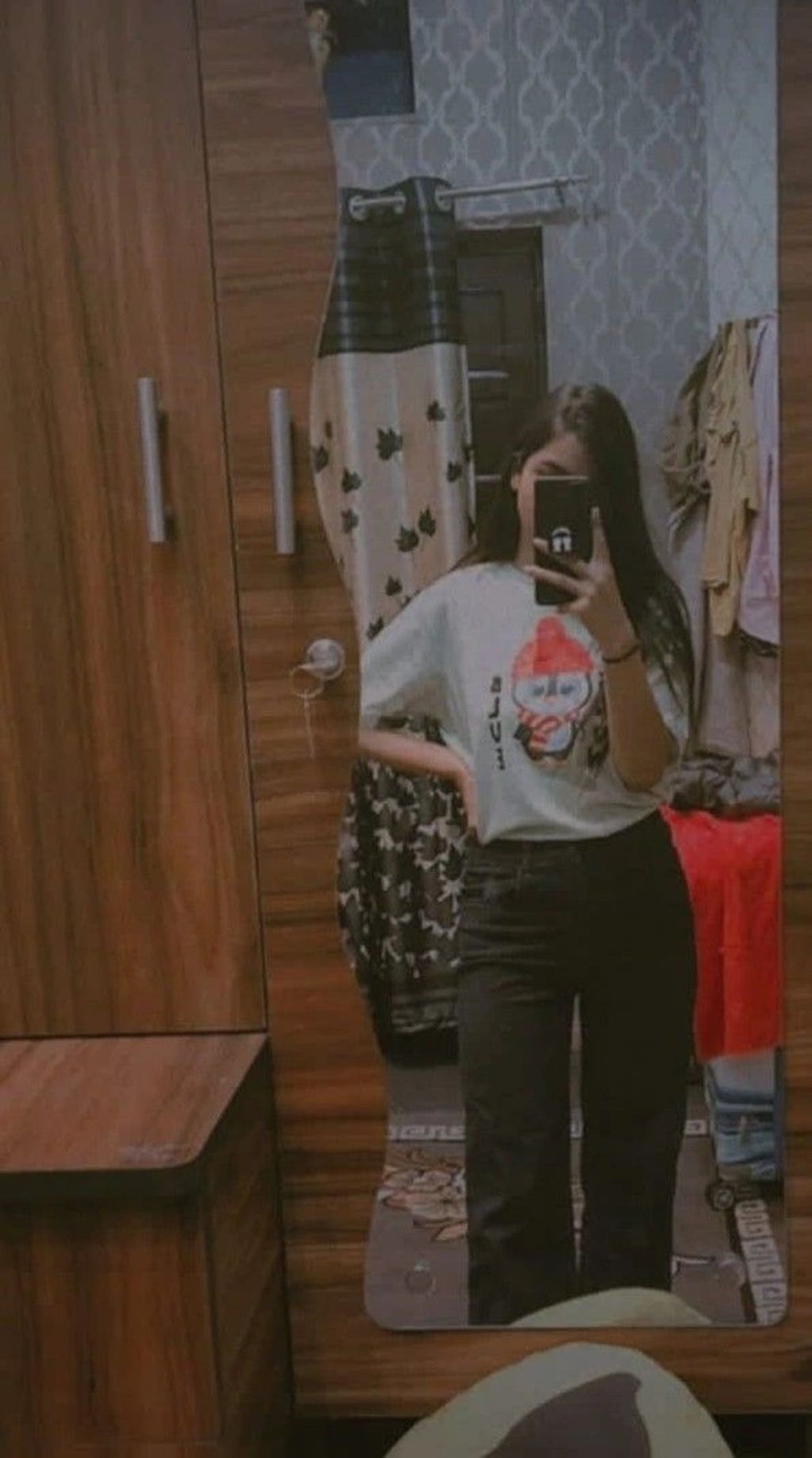 A person takes a mirror selfie in a room with wooden furniture and patterned wallpaper.