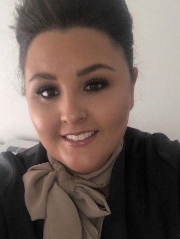 A smiling woman with dark hair and makeup wearing a black outfit with a beige bow.