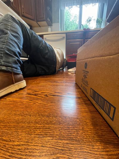 Dishwasher repairman laying on floor reaching under GE dishwasher next to a GE dishwasher part box