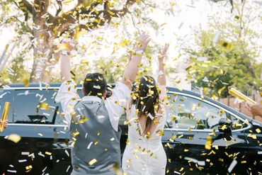 Newlyweds celebrating with gold and white confetti outside a black car.