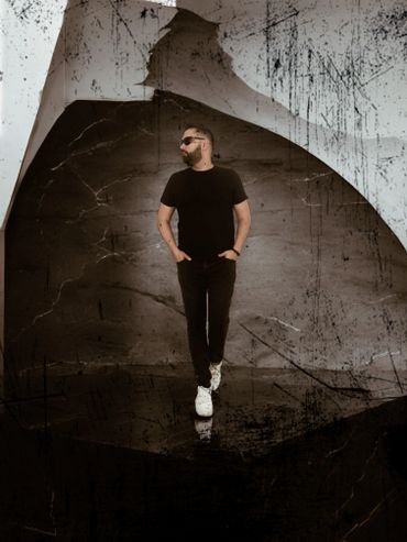 Man in black outfit and white shoes standing against a textured dark background.