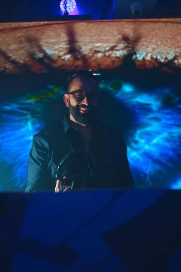 Man with headphones stands before a giant eye projection in a dark room.