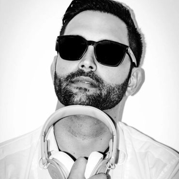 Man with beard and sunglasses holding headphones, posing confidently.