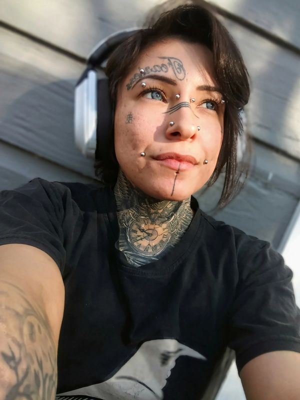 Person with facial tattoos and piercings wearing headphones and a black shirt.