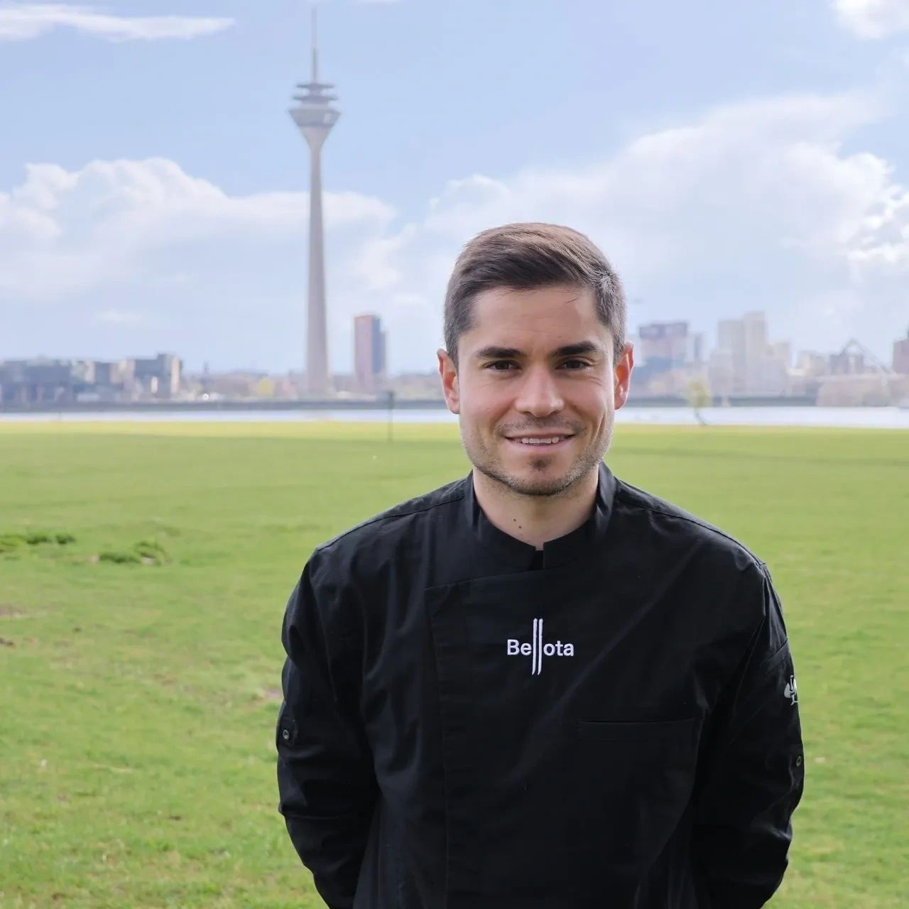 Smiling chef in black uniform outdoors with cityscape and tower in background.