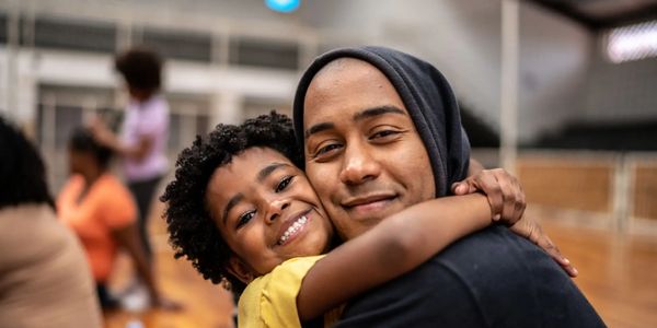 A joyful child hugging an adult wearing a hoodie in a gym setting.
