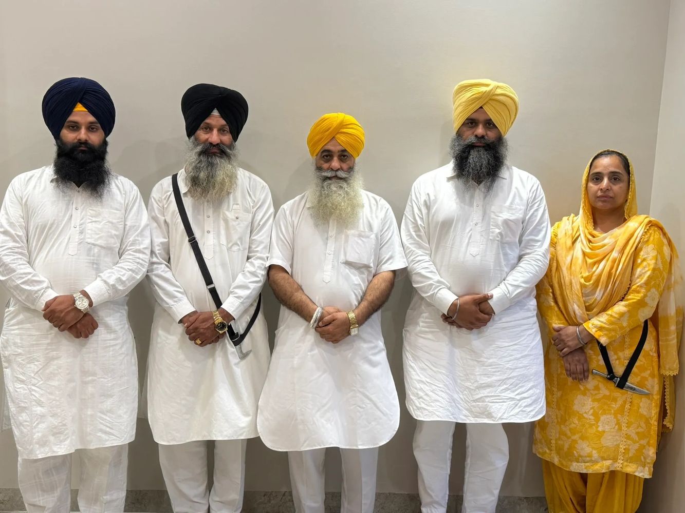 Group of Sikh men and a woman in traditional attire standing against a plain wall.