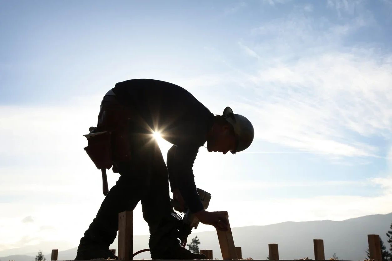 Silhouetted construction worker fixing wood under a bright sky.