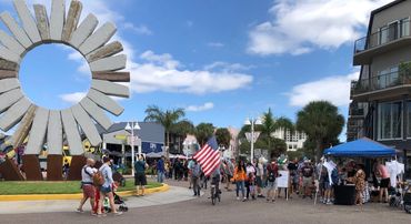 People gather at a sunny outdoor event near a distinctive circular sculpture.