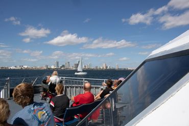 People on a boat enjoying a sunny day with a city skyline and sailboat in the background.