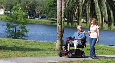 A man on a mobility scooter and a woman walking beside him by a lakeside path.