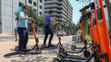 Two men preparing to ride orange electric scooters in an urban setting.