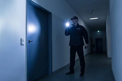 Security guard inspecting a door with a flashlight in a dark hallway.