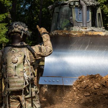 Soldier signals a bulldozer in a wooded area during military operations.