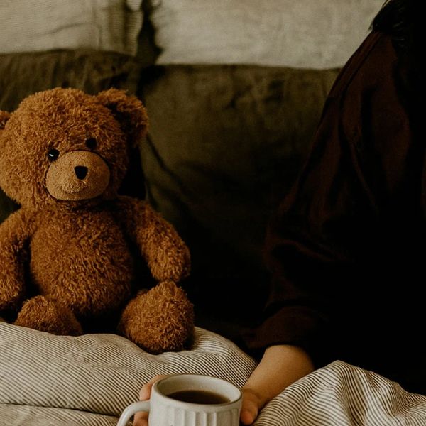A teddy bear sits on a cozy bed next to a person holding a cup of coffee.