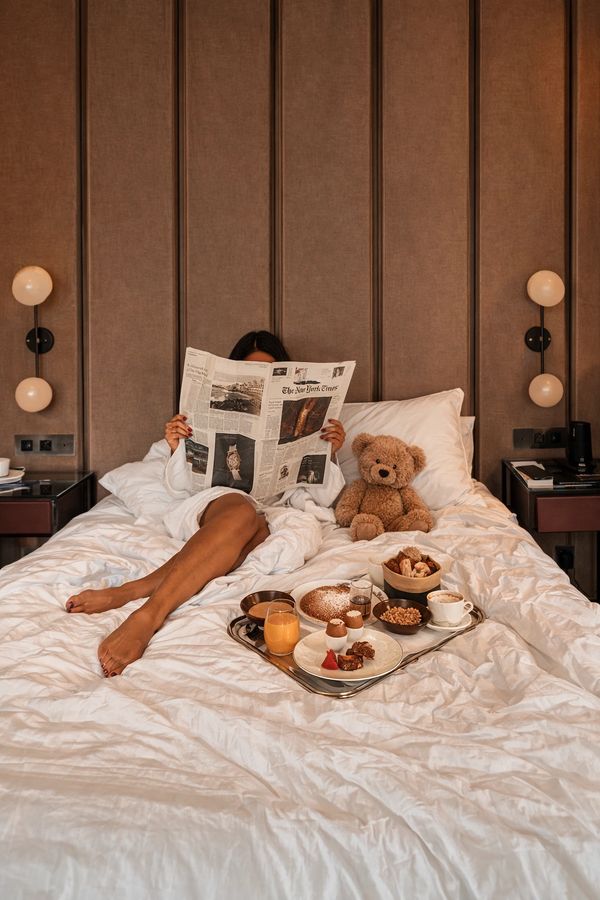 Person in bed reading newspaper with teddy bear and breakfast tray.