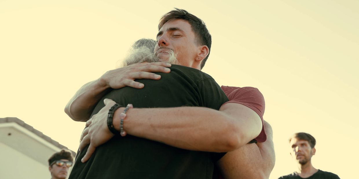 Two men sharing a heartfelt hug outdoors with two others watching in the background.