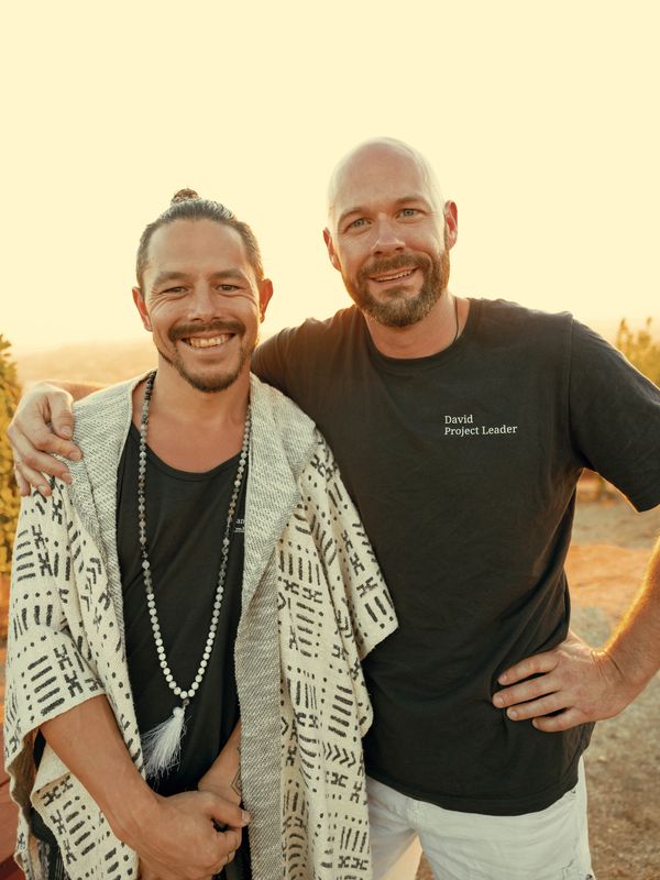 Two men smiling outdoors during sunset, one wearing a patterned shawl and the other a shirt labeled 'David Project Leader'.